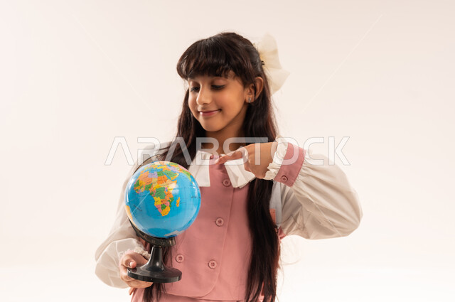 Back to school, preparing for the new school year, the concept of research and exploration around the world, a close-up portrait of an Arab Gulf Omani student wearing a school uniform holding a globe, a Saudi girl looking with gestures of happiness and pleasure, white background
