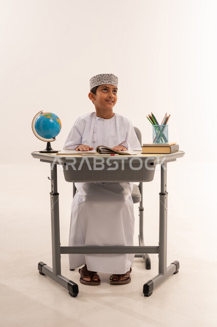 متابعة المناهج الدراسية بمثابرة وعزيمة ، مفهوم البحث والإستكشاف حول العالم، بورتريه لطالب عربى خليجي عماني يرتدي الدشداشة و الكمه يجلس على طاوله مكتبية يقوم بمذاكره الدروس المدرسيه و ينظر الى شيء ما، خلفية بيضاء
