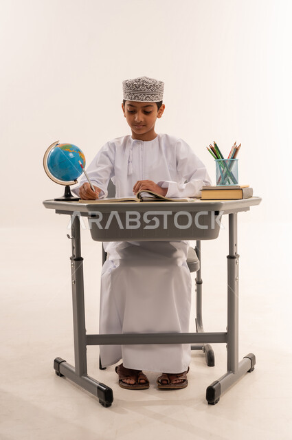 مفهوم الاجتهاد وطلب التفوق، المقررات والمناهج التعليمية الحديثة والمطورة، موسم العودة للمدارس، بورتريه لطالب عربى خليجي عماني يرتدي الدشداشة و الكمه يجلس على طاوله مكتبية يقوم  بمذاكره الدروس المدرسيه، خلفية بيضاء