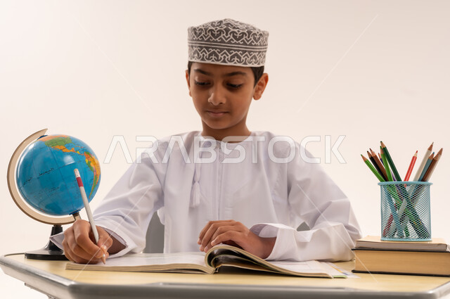 المقررات والمناهج التعليمية الحديثة والمطورة، مفهوم الاجتهاد وطلب التفوق، موسم العودة للمدارس، بورتريه مقرب لطالب عربى خليجي عماني يرتدي الدشداشة و الكمه يجلس على طاوله مكتبية يقوم  بمذاكره الدروس المدرسيه، خلفية بيضاء