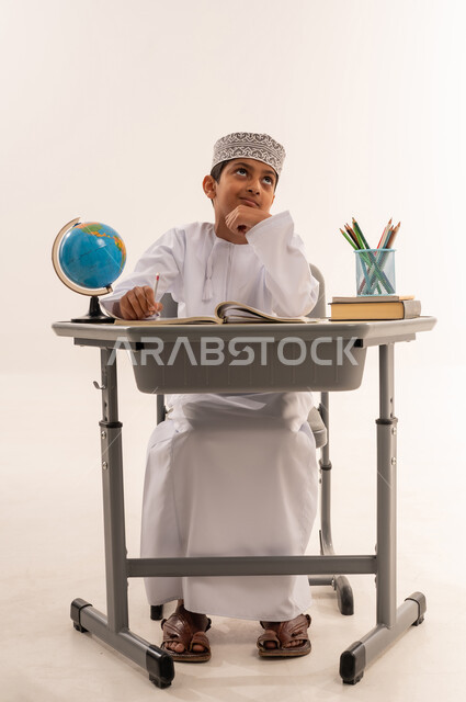 الإستعداد للعام الدراسي الجديد، تأدية الواجبات المدرسية، بورتريه لطالب عربي خليجي عماني يرتدي الدشداشة و الكمة يجلس على الطاولة بجانبه مجموعة من الكتب المدرسية ومجسم الكرة الأرضية و يقوم بالتفكير فى شي ما، خلفية بيضاء