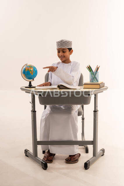 تطوير المناهج التعليمية في سلطنه عمان، مفهوم البحث والإستكشاف حول العالم، بورتريه لطالب عربى خليجي عماني الزي التقليدي يجلس على طاوله مكتبية يقوم بالاشارة الى مجسم الكرة الأرضية، مذاكره الدروس المدرسيه، خلفية بيضاء