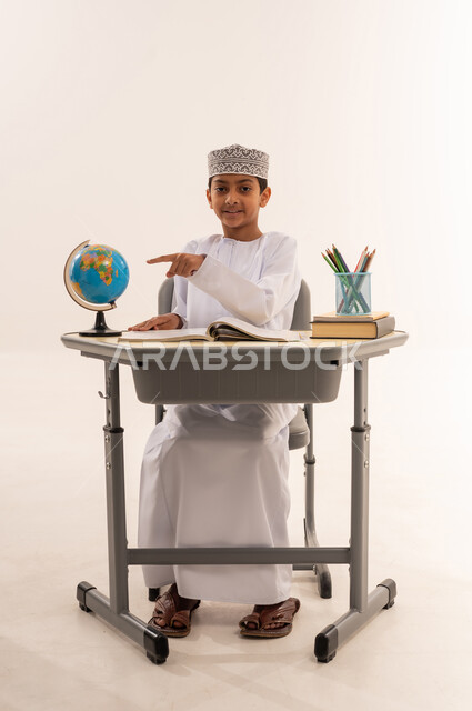 مفهوم البحث والإستكشاف حول العالم، جودة أساليب التعلم الفعال وباستخدام الأدوات والوسائل المساعدة، بورتريه لطالب عربى خليجي عماني الزي التقليدي يجلس على طاوله مكتبية يقوم بالاشارة الى مجسم الكرة الأرضية، مذاكره الدروس المدرسيه، خلفية بيضاء
