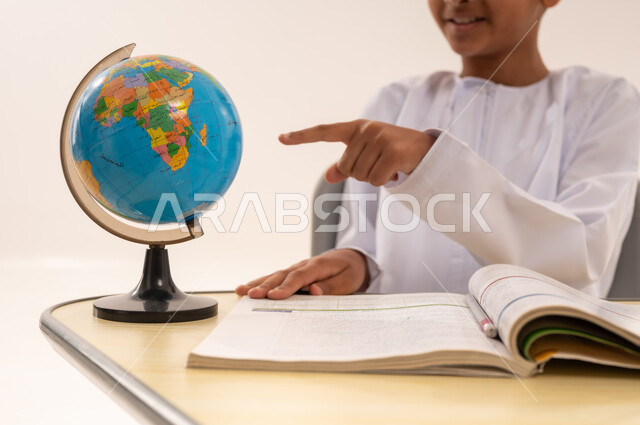 جودة أساليب التعلم الفعال وباستخدام الأدوات والوسائل المساعدة، مفهوم البحث والإستكشاف حول العالم، بورتريه مقرب لطالب عربى خليجي عماني الزي التقليدي يجلس على طاوله مكتبية يقوم بالاشارة الى مجسم الكرة الأرضية، مذاكره الدروس المدرسيه، خلفية بيضاء