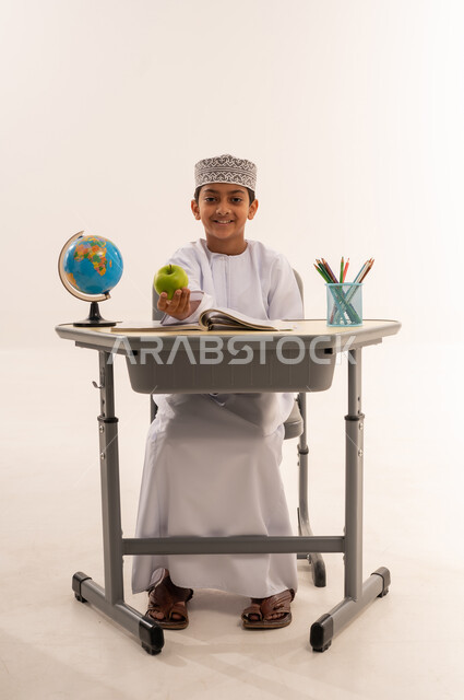 الاجتهاد للوصول إلى النجاح والتفوق، موسم العودة للمدارس، بورتريه لطالب عربي خليجي عماني يرتدي الدشداشة و الكمه يجلس على الطاوله المكتبية و يقوم بالنظر إلى الكاميرا بإيماءات الاندماج والسعادة يمسك بيده تفاحه خضراء ، حل الواجبات والمراجعة، خلفية بيضاء