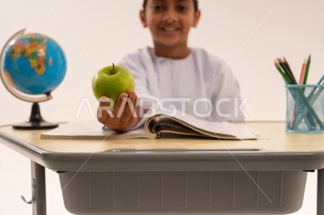 Back to school season, hard work to achieve success and excellence, close-up portrait of an Arab Gulf Omani student wearing a dishdasha and a turban sitting at an office table and looking at the camera with gestures of integration and happiness holding a green apple in his hand, doing homework and reviewing, white background