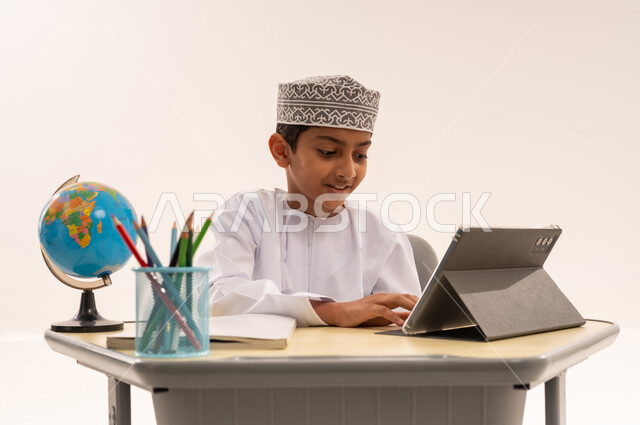 مفهوم التعلم عبر الانترنت، مراجعة الدروس اليوميه، ادخال التقنية والتكنولوجيا بالتعليم، بورتريه مقرب لطالب عربى خليجي عماني مبتسم يرتدي الدشداشة و الكمه يجلس على الطاولة المكتبيه يقوم بحل الواجبات وتلقي المحاضرات المدرسيه عن بعد عبر التابلت، خلفية بيضاء