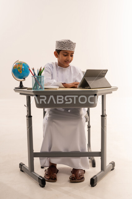 مفهوم العودة إلى المدارس, مراجعة الدروس اليوميه، ادخال التقنية والتكنولوجيا بالتعليم، بورتريه لطالب عربى خليجي عماني مبتسم يرتدي الدشداشة و الكمه يجلس على الطاولة المكتبيه يقوم بحل الواجبات وتلقي المحاضرات المدرسيه عن بعد عبر التابلت، خلفية بيضاء