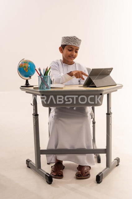 مراجعة الدروس اليوميه، ادخال التقنية والتكنولوجيا بالتعليم، الفصل الدراسي جديد، بورتريه لطالب عربى خليجي عماني مبتسم يرتدي الدشداشة و الكمه يجلس على الطاولة المكتبيه يقوم بحل الواجبات وتلقي المحاضرات المدرسيه عن بعد عبر التابلت، خلفية بيضاء