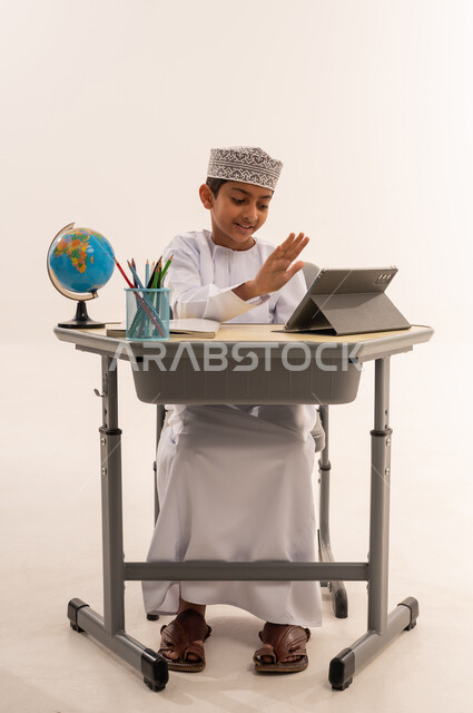 ادخال التقنية والتكنولوجيا بالتعليم، الفصل الدراسي جديد، مراجعة الدروس اليوميه، بورتريه لطالب عربى خليجي عماني مبتسم يرتدي الدشداشة و الكمه يجلس على الطاولة المكتبيه يقوم بحل الواجبات وتلقي المحاضرات المدرسيه عن بعد عبر التابلت، خلفية بيضاء