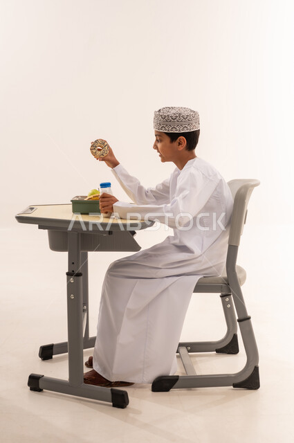 العناصر الغذائية الهامة لبناء الجسم، الاستمتاع بتناول الأطعمة الصحية في وقت الفسحة المدرسية، بورترية  من الجانب لطالب عربى خليجي عماني يرتدي الدشداشة و الكمه يجلس على الطاوله المكتبية يقوم بتناول قطعه من المخبوزات ، خلفية بيضاء