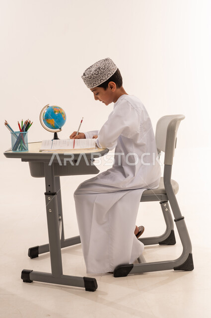 مراجعة الدروس اليومية ، ايماءات تدل على التركيز والتفكير بالإجابة، طلب العلا والمثابرة والحرص على النجاح، بورتريه من الجانب لطاب عربي خليجي عماني يرتدي الدشداشة و الكمة يجلس على طاولة مكتبية يقوم بالمذاكرة للاختبارات والاستعداد للامتحان، خلفية بيضاء