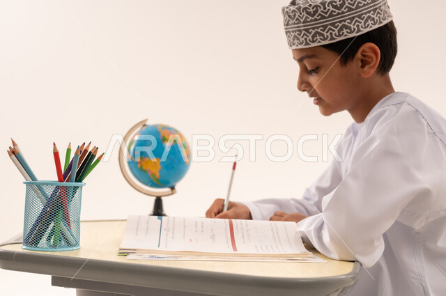 Gestures indicating concentration and thinking about the answer, seeking excellence, perseverance and keenness to succeed, close-up side portrait of an Arab Gulf Omani student wearing a dishdasha and a kummah sitting at an office table studying for tests and preparing for the exam, white background