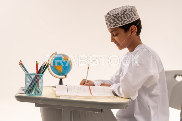 طلب العلا والمثابرة والحرص على النجاح، ايماءات تدل على التركيز والتفكير بالإجابة، بورتريه مقرب من الجانب لطاب عربي خليجي عماني يرتدي الدشداشة و الكمة يجلس على طاولة مكتبية، المذاكرة للاختبارات والاستعداد للامتحان، خلفية بيضاء