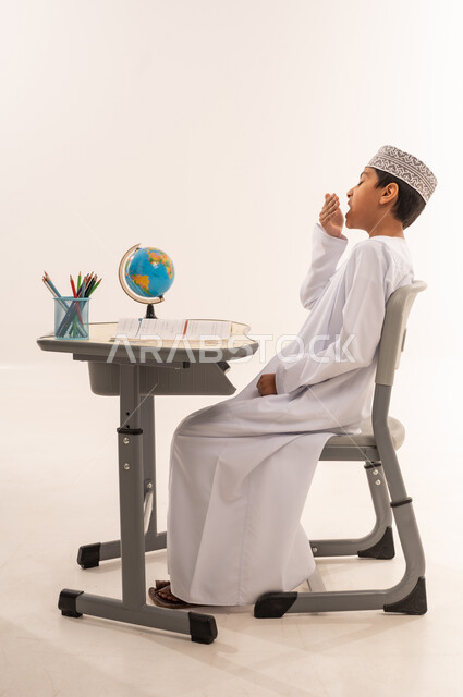 الإحساس بالتعب والتثاؤب، الشعور بالكسل والخمول، العودة إلى المدارس، بورترية من الجانب لطالب عربي خليجي عماني يرتدي الدشداشة و الكمه يجلس على الطاوله المكتبية يرفع يده على فمه بإيماءات النعاس،الحاجه إلى الراحه, الحاجه الى القيلوله, خلفية بيضاء