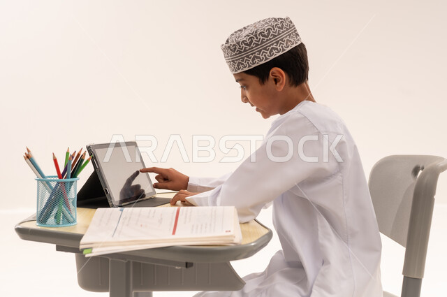Integrating modern technology devices into learning, reviewing daily lessons, new semester, close-up side portrait of an Arab Gulf Omani student wearing a dishdasha and a keffiyeh sitting at an office table doing homework and receiving school lectures remotely via tablet, white background