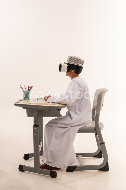 جمع التعلم مع الترفيه بإستخدام التقنيه والتكنولوجيا الحديثه، بورتريه من الجانب لطالب عربي خليجي عماني يرتدي الدشداشة و الكمة و نظارة الواقع الإفتراضي ثلاثية الأبعاد يجلس على طاوله مكتبيه يستمتع بمشاهده فيلم، خلفية بيضاء