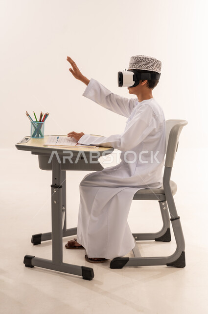 محاكاة العالم الافتراضي ، بورتريه من الجانب لطالب عربي خليجي عماني يرتدي الدشداشة و الكمة و نظارة الواقع الإفتراضي ثلاثية الأبعاد يجلس على طاوله مكتبيه يلعب بإيماءات و تعابير يدين تدل على الاندماج، خلفية بيضاء
