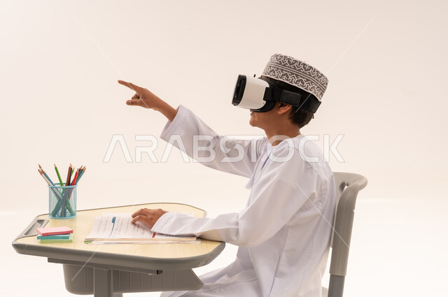 اتباع طرق تعليمية جديدة، دمج التقنيات الحديثة في الحياة العلمية، اكاديميات ومدارس متطورة، بورتريه مقرب من الجانب لتلميذ عربى خليجي عمانى يرتدي الدشداشة و الكمه يجلس على طاوله مكتبية و يقوم بالاشاره الى شيء ما، الاستمتاع بالمشاهده عبر نظارة الواقع الافتراضي ، خلفية بيضاء