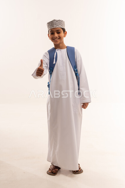 موسم العودة إلى المدارس، الجد والاجتهاد للوصول إلى النجاح والتفوق، بورتريه لطالب عربي خليجي عماني يرتدي الدشداشة و الكمة يحمل حقيبة الظهر المدرسية يقوم برفع اصبع الابهام للاعلى بايماءات الموافقة والقبول، الجسم بالطول الكامل، خلفية بيضاء