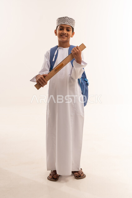 العودة إلى المدارس، بورتريه لطالب عربي خليجي عماني مبتسم يرتدي الدشداشة و الكمه يحمل حقيبة الظهر المدرسية وبيده مسطرة مرقمة يقوم بالنظرالى الكاميرا بايماءات السعادة و السرور، التعليم الدراسي في سلطنه عمان، الجسم بالطول الكامل، خلفية بيضاء