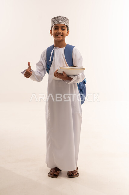 Thumbs up gesture of approval and acceptance, back to school season, hard work and diligence to achieve success and excellence, portrait of an Arab Gulf Omani student wearing a dishdasha and a kummah carrying a school backpack, standing and holding books and scientific curricula, full body, white background