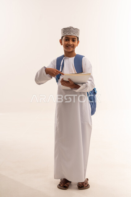 موسم العودة إلى المدارس، الجد والاجتهاد للوصول إلى النجاح والتفوق، بورتريه لطالب عربي خليجي عماني يرتدي الدشداشة و الكمة يحمل حقيبة الظهر المدرسية يقف ويمسك بالكتب والمناهج العلمية و يقوم بالاشارة اليها، الجسم بالطول الكامل، خلفية بيضاء