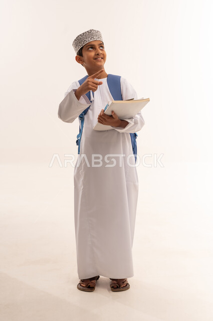 الجد والاجتهاد للوصول إلى النجاح والتفوق، موسم العودة إلى المدارس، بورتريه لطالب عربي خليجي عماني يرتدي الدشداشة و الكمة يحمل حقيبة الظهر المدرسية يقف ويمسك بالكتب والمناهج العلمية و يقوم بالاشارة و النظر الى شيء ما، الجسم بالطول الكامل، خلفية بيضاء