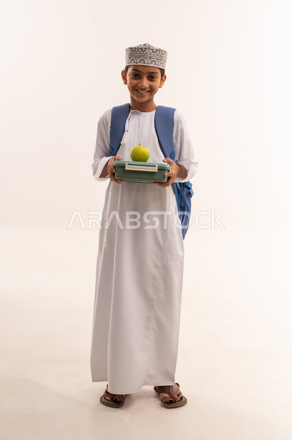 الفطور الصباحي الصحي و المفيد، الاستعداد للعام الدراسي الجديد, بورتريه لطالب عربي خليجي عماني مبتسم يرتدي الدشداشه و الكمه و الحقيبه المدرسية يحمل بيده تفاحة خضراء و لانش بوكس، الجسم بالطول الكامل، خلفية بيضاء