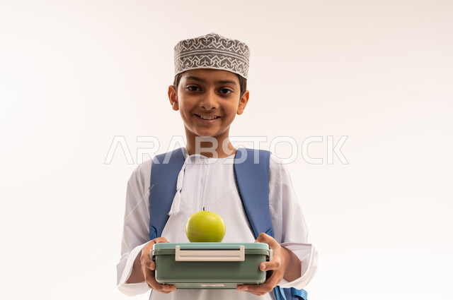 تناول الأطعمة الصحية في وقت الفسحة المدرسية، الاستعداد للعام الدراسي الجديد, بورتريه مقرب لطالب عربي خليجي عماني مبتسم يرتدي الدشداشه و الكمه و الحقيبه المدرسية يحمل بيده تفاحة خضراء و لانش بوكس، خلفية بيضاء