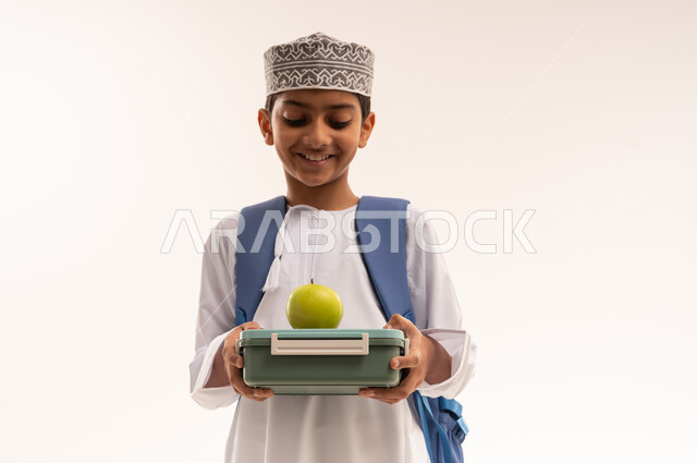 الاستعداد للعام الدراسي الجديد، أهمية تناول الطعام الصحي والمتوازن للحفاظ على صحة الجسم والعقل, بورتريه مقرب لطالب عربية خليجي عماني مبتسم يرتدي الدشداشه و الكمه و الحقيبه المدرسية يحمل بيده تفاحة خضراء و لانش بوكس، خلفية بيضاء