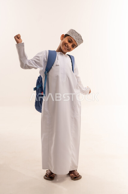 مفهوم النشاط والإستيقاظ الصباحي، بورتريه لطالب عربي خليجي عماني يرتدي الدشداشة و الكمه و الحقيبه المدرسيه يرفع يديه بإيماءات تدل على التمدد ، الإستعداد للعام الدراسي الجديد، التعليم الدراسي في سلطنه عمان، الجسم بالطول الكامل، خلفية بيضاء