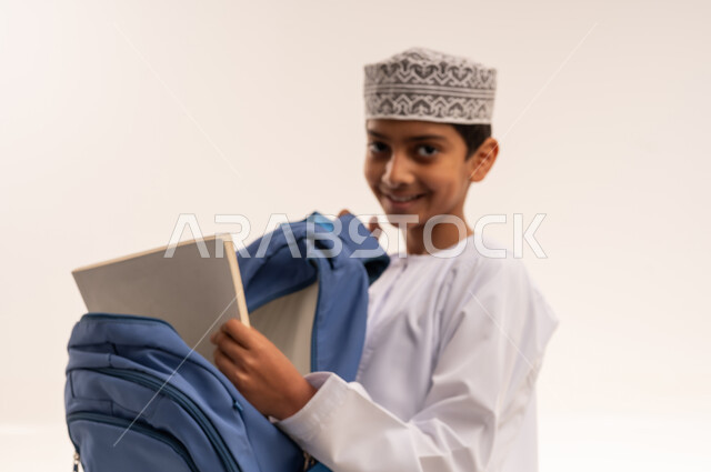 الاستعداد للذهاب الى المدرسة، بورترية مقرب لطالب عربي خليجي عماني يرتدي الدشداشة والكمه يقوم بتجهيز وتنظيم الحقيبة المدرسية، سنه دراسية و فصل دراسي جديد، المناهج العلمية التعليمية، خلفية بيضاء