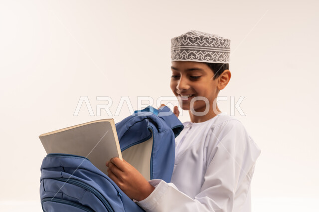 سنه دراسية و فصل دراسي جديد، الاستعداد للذهاب الى المدرسة، بورترية مقرب لطالب عربي خليجي عماني يرتدي الدشداشة والكمه يقوم بتجهيز وتنظيم الحقيبة المدرسية، المناهج العلمية التعليمية، خلفية بيضاء