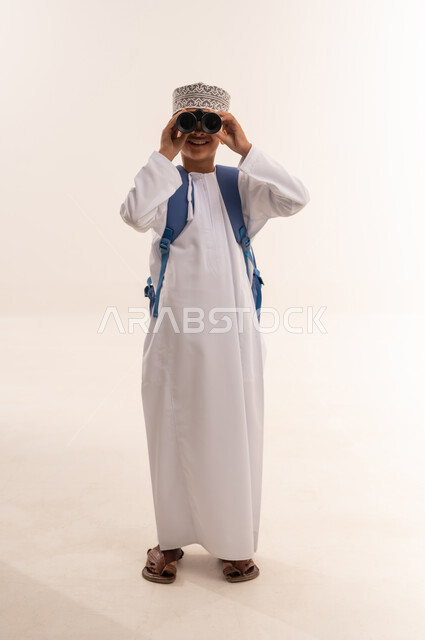 تقريب الأشياء والأماكن البعيدة، حب الاستكشاف و المغامرة, التخطيط والأهداف المستقبلية، توضيح الرؤية عبر التلسكوب, بورتريه لطالب عربي خليجي عماني يرتدي الدشداشة والكمه و الحقيبه المدرسية يحمل بيده منظار ثنائي العينين و ينظر الى شيء ما, الجسم بالطول الكامل، خلفية بيضاء