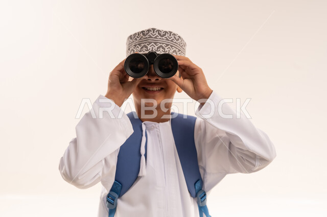 التخطيط والأهداف المستقبلية، حب الاستكشاف و المغامرة، تقريب الأشياء والأماكن البعيدة، توضيح الرؤية عبر التلسكوب, بورتريه مقرب لطالب عربي خليجي عماني يرتدي الدشداشة والكمه و الحقيبه المدرسية يحمل بيده منظار ثنائي العينين و ينظر الى شيء ما، خلفية بيضاء