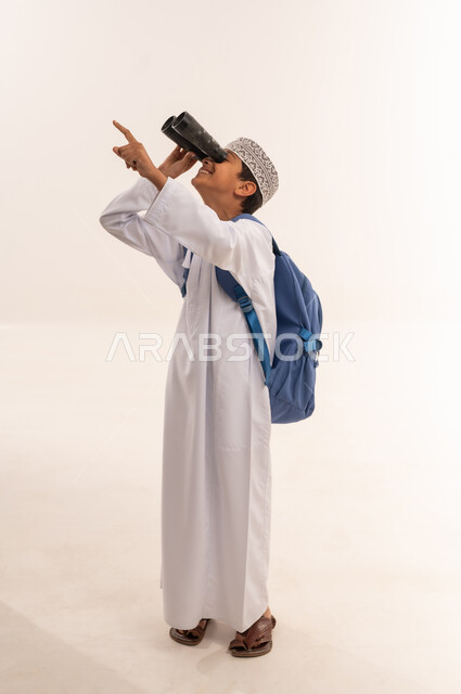 حب الاستكشاف و المغامرة, التخطيط والأهداف المستقبلية، تقريب الأشياء والأماكن البعيدة، توضيح الرؤية عبر التلسكوب, بورتريه لطالب عربي خليجي عماني يرتدي الدشداشة والكمه و الحقيبة المدرسية يحمل بيده منظار ثنائي العينين و ينظر الى شيء ما, الجسم بالطول الكامل، خلفية بيضاء