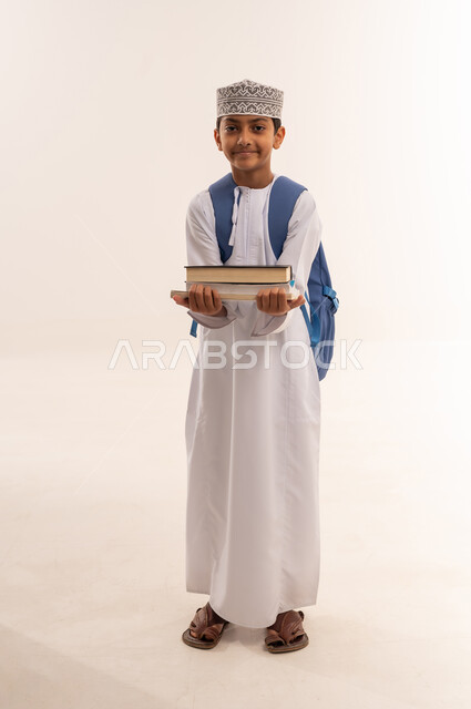 Reviewing lessons and solving homework, back to school, the concept of seriousness and diligence to achieve success, educational scientific curricula, portrait of an Arab Gulf Omani student wearing a dishdasha and a kuma carrying a school backpack and holding a group of books, a full body image, white background