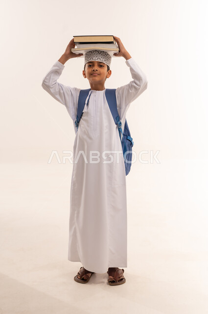 العودة الى المدارس، مفهوم الجد والاجتهاد للوصول إلى النجاح، المناهج العلمية التعليمية، مراجعة الدروس وحل الواجبات، بورتريه لطالب عربي خليجي عماني يرتدي الدشداشة والكمة يحمل حقيبة الظهر المدرسية يحمل بيده مجموعة من الكتب على رأسة، صورة لكامل الجسم، خلفية بيضاء