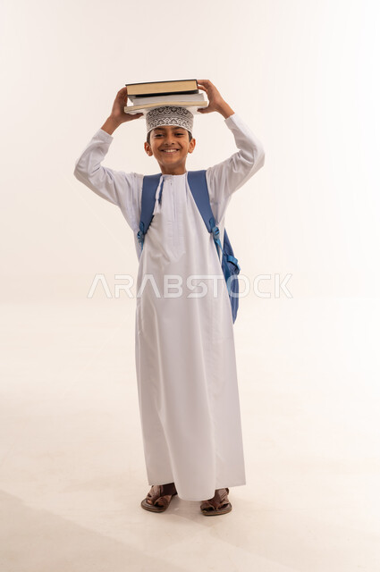 مفهوم الجد والاجتهاد للوصول إلى النجاح، المناهج العلمية التعليمية، مراجعة الدروس وحل الواجبات، العودة الى المدارس، بورتريه لطالب عربي خليجي عماني يرتدي الدشداشة والكمة يحمل حقيبة الظهر المدرسية يحمل بيده مجموعة من الكتب على رأسة، صورة لكامل الجسم، خلفية بيضاء