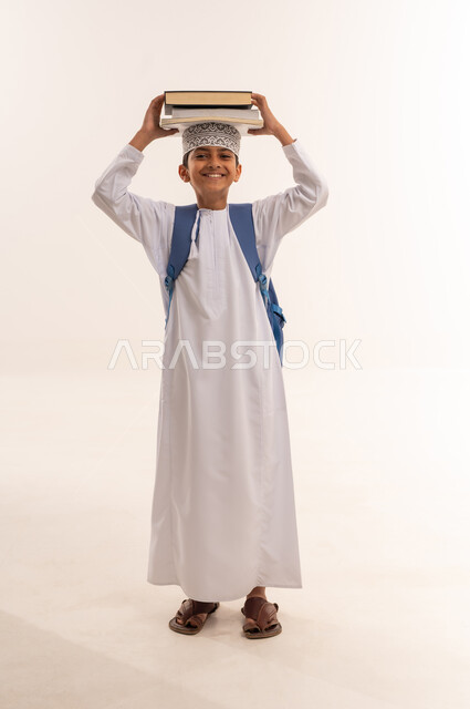 المناهج العلمية التعليمية، مفهوم الجد والاجتهاد للوصول إلى النجاح، مراجعة الدروس وحل الواجبات، العودة الى المدارس، بورتريه لطالب عربي خليجي عماني يرتدي الدشداشة والكمة يحمل حقيبة الظهر المدرسية يمسك بيده مجموعة من الكتب، صورة لكامل الجسم، خلفية بيضاء