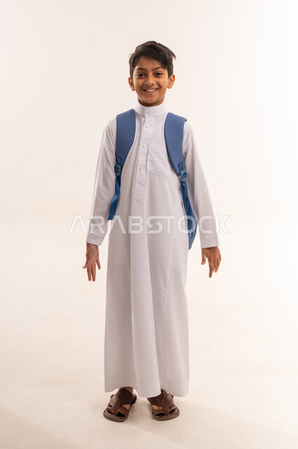 المناهج العلمية التعليمية، مفهوم الاجتهاد للوصول إلى النجاح، مفهوم العودة إلى المدارس، بورتريه لطالب عربي خليجي عماني يرتدي الثوب التقليدي يحمل حقيبة الظهر المدرسية ، ولد سعودي يستعد للذهاب الى المدرسة، صورة لكامل الجسم، خلفية بيضاء