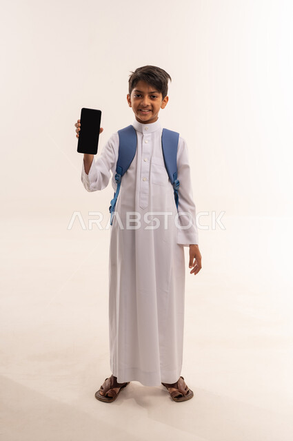 استخدام الأجهزة التقنية الحديثة, بورتريه لفتى عربي خليجي عماني مبتسم يرتدي الثوب التقليدي ويحمل الحقيبة المدرسية يقوم بعرض شاشة سوداء فارغة عبر الهاتف المحمول, ولد سعودي ينظر بايماءات السعاده و السرور,الجسم بالطول الكامل، خلفية بيضاء