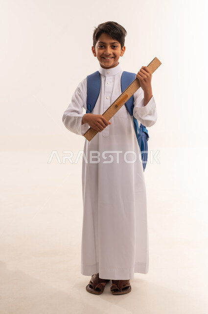 العودة إلى المدارس، بورتريه لطالب عربي خليجي عماني مبتسم يرتدي الزي التقليدي يحمل حقيبة الظهر المدرسية وبيده مسطرة مرقمة، أدوات قرطاسية، ولد سعودي ينظر الى الكاميرا بايماءات السعادة و السرور ، التعليم الدراسي في سلطنه عمان، الجسم بالطول الكامل، خلفية بيضاء