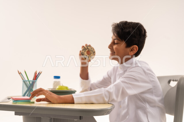 الوجبات الغنية بالعناصر الغذائية الهامة، موسم العودة إلى المدارس، ولد سعودي ينظر بإيماءات السعادة والسرور، القرطاسية المدرسية والأقلام، بورتريه مقرب من الجانب لطالب عربي خليجي عماني يرتدي الثوب التقليدي يجلس في مقعده يمسك بيده قطعه من المخبوزات، خلفية بيضاء