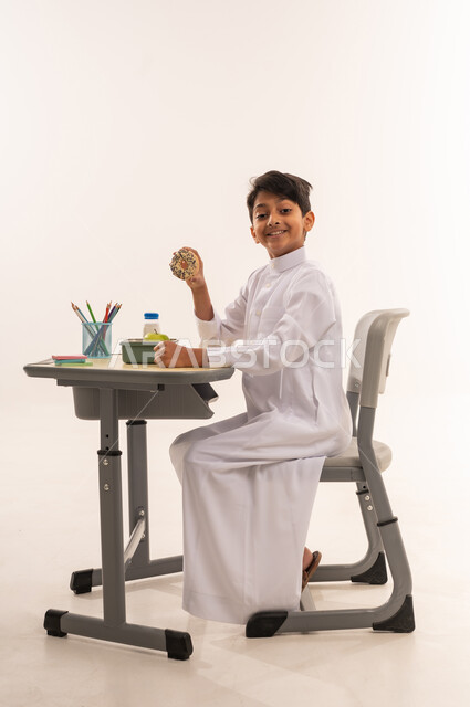 لانش بوكس يحتوي على سناكات صحية مغذية، موسم العودة إلى المدارس، ولد سعودي ينظر بإيماءات السعادة والسرور، القرطاسية المدرسية والأقلام، بورتريه من الجانب لطالب عربي خليجي عماني يرتدي الثوب التقليدي يجلس في مقعده يمسك بيده قطعه من المخبوزات، خلفية بيضاء