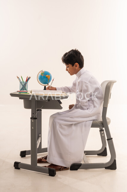 Focus and integration during study, beginning of a new school year, goals of education, portrait of an Arab Gulf Omani student wearing traditional dress sitting at a table studying, Saudi boy working hard to write exercises, quality of education in schools of the Sultanate of Oman, white background