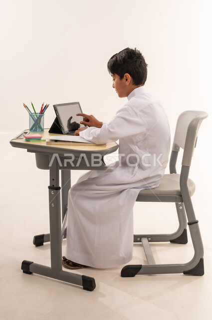دمج الأجهزة التقنية الحديثة بالتعلم، الفصل الدراسي جديد، بورتريه من الجانب لتلميذ عربى خليجي عماني يرتدي الزي التقليدي يجلس على الطاولة المكتبيه ، ولد سعودي يقوم بحل الواجبات وتلقي المحاضرات المدرسيه عن بعد عبر التابلت، خلفية بيضاء