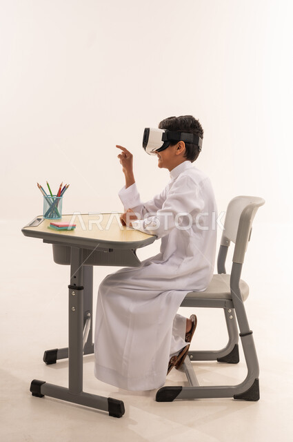 Development of teaching methods using modern advanced technologies, a Saudi student sitting at a study table, advanced academies and schools, a side portrait of an Arab Gulf Omani student wearing traditional clothing enjoying watching through virtual reality glasses, white background