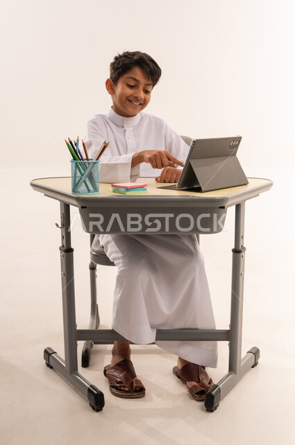متابعة الحصص و حل الواجبات عبر الانترنت ،ولد سعودى يقوم بمتابعة الدروس عن طريق منصات التعلم عن بعد، بورتريه لطالب عربي سعودي خليج يرتدي الزي الرسمي يجلس أمام طاولة الدراسة والتابلت، إيماءات التفاعل والمشاركة، خلفية بيضاء